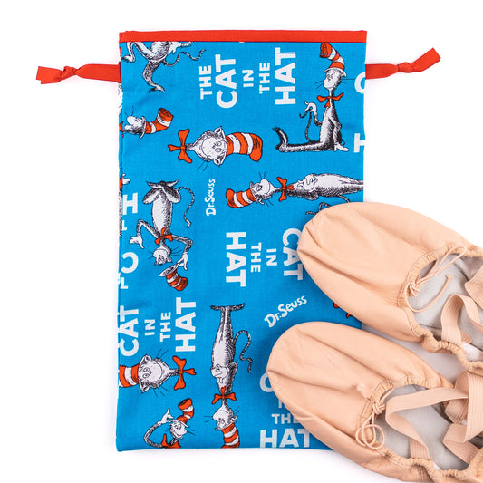 A pair of pink flat-soled ballet dance shoes next to a blue, red, and white cat in the hat themed shoe bag