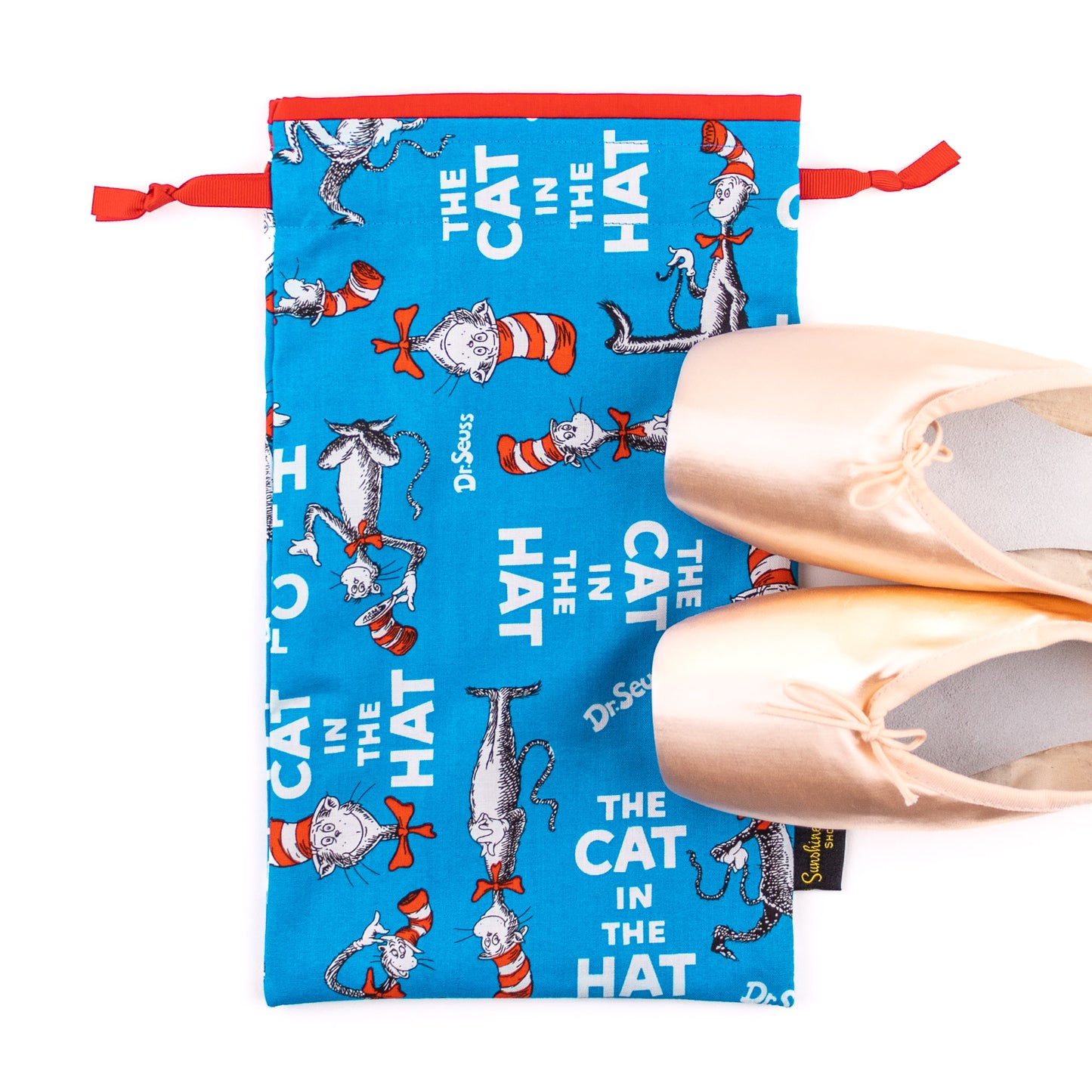 A pair of pink pointe ballet dance shoes next to a blue, red, and white "Cat in the Hat" themed shoe bag